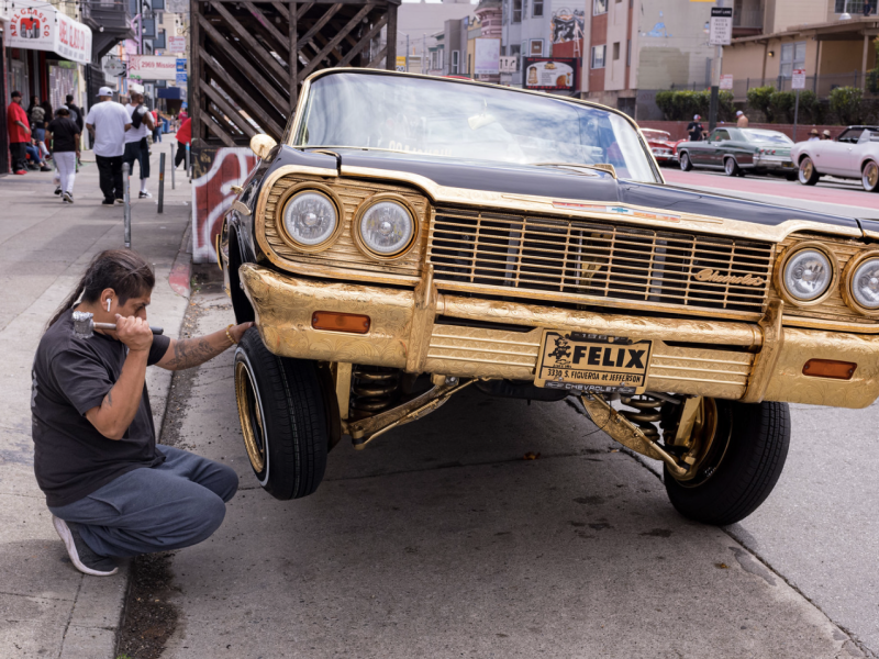 Man squats next to a gold-trimmed car with raised hydraulics on a city street while talking on a phone.